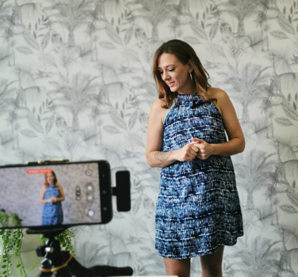 A woman in a blue patterned dress stands in front of a camera, recording content against a leafy wallpaper background.